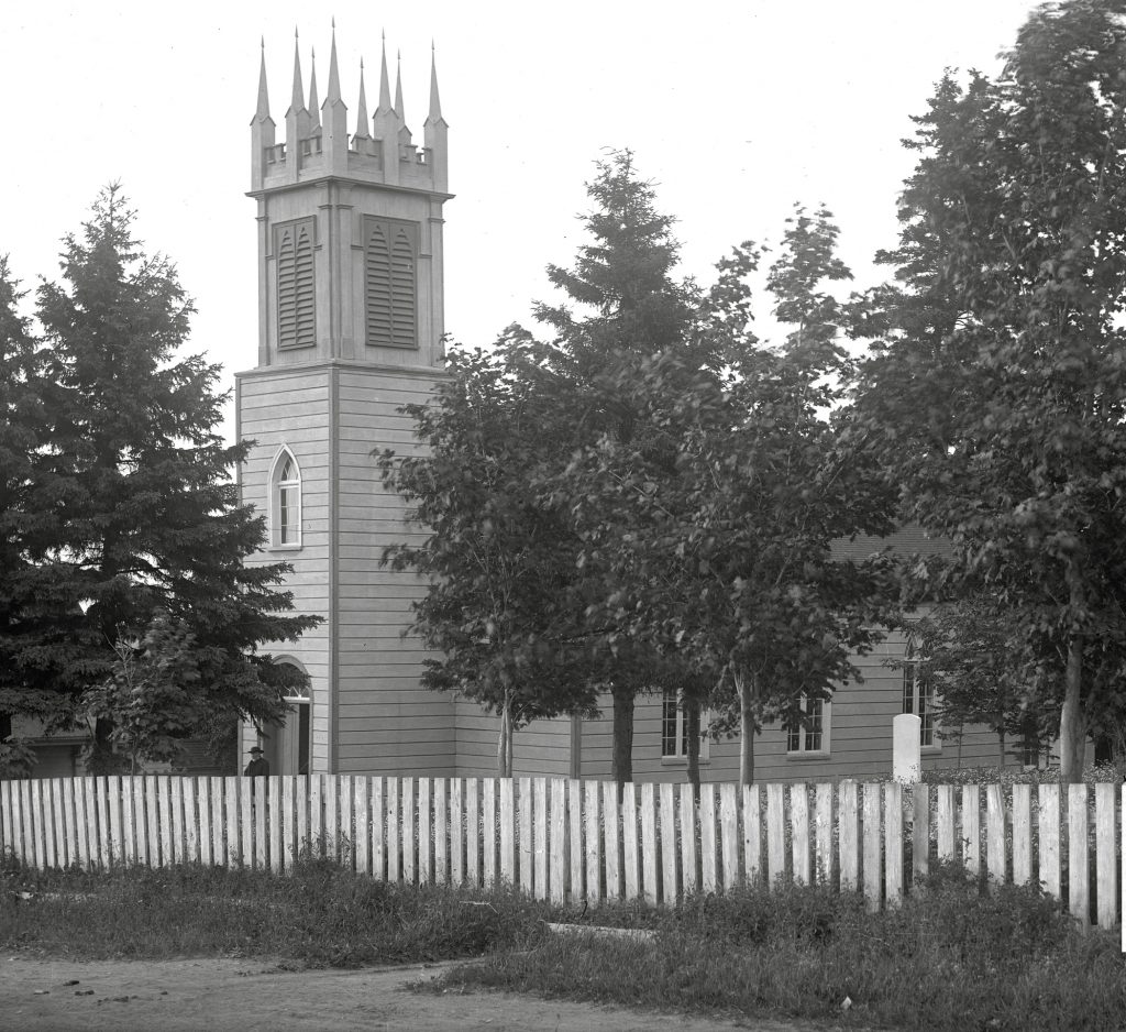 Photo noir et blanc d'une vieille église en bois, église anglicane St. Bartholomew, surmontée d’un clocher en façade; un prêtre sortant de son entrée principale. Au premier plan, une clôture en bois sur le fronton de l'église, derrière laquelle se dressent de grands arbres.