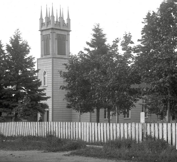 Black and white photo of an old wooden church, St. Bartholomew's Anglican church with a steeple in front, one person emerging from the front entrance, and a picket fence all across the front of the church property, in the foreground of the photo. Also there are several large trees between the fence and the church.