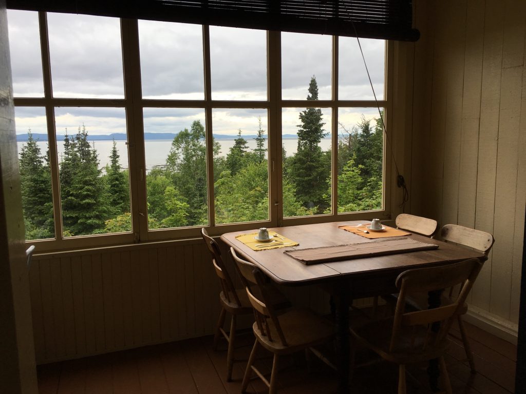 A colour photo of a small room featuring three sets of quarter-paned windows that reveal woods, river, and sky. Inside the small room, on the viewer's side of the windows, is a small wooden table with five wooden chairs and breakfast places set for two people.