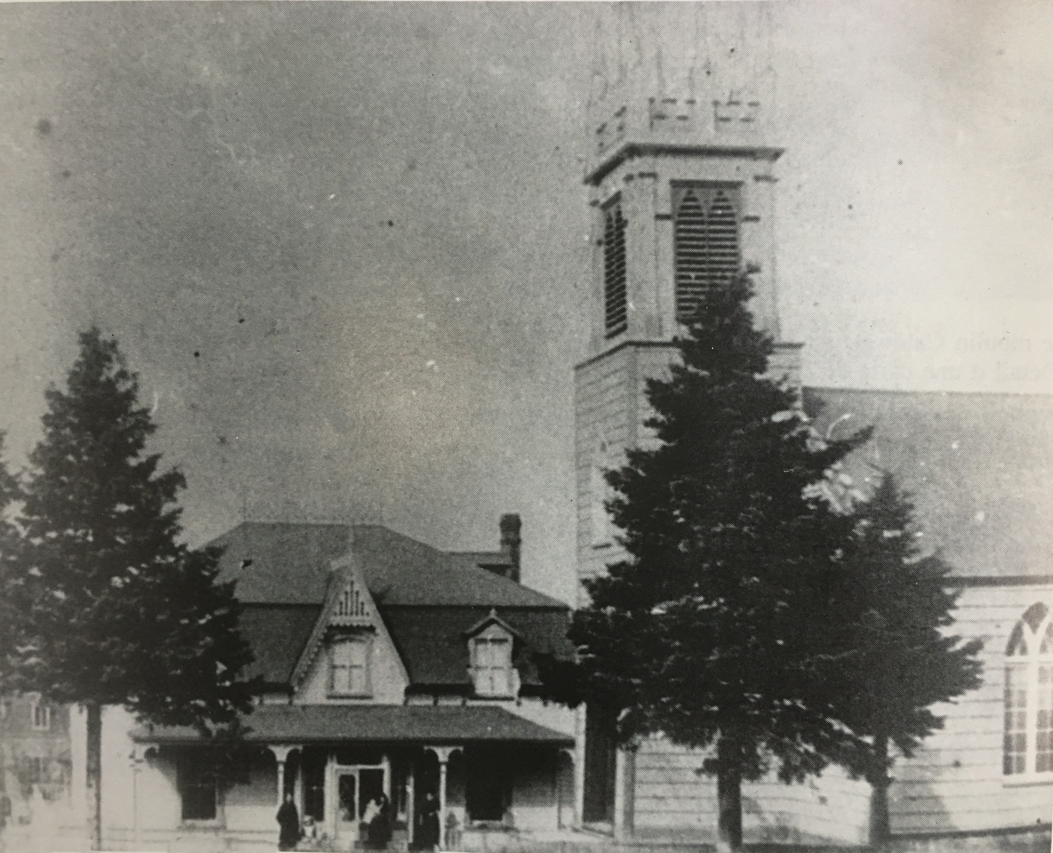 A black and white photograph of an old wooden church with a steeple at the front, and a two-storey minister's house erected quite close to the front of the church.