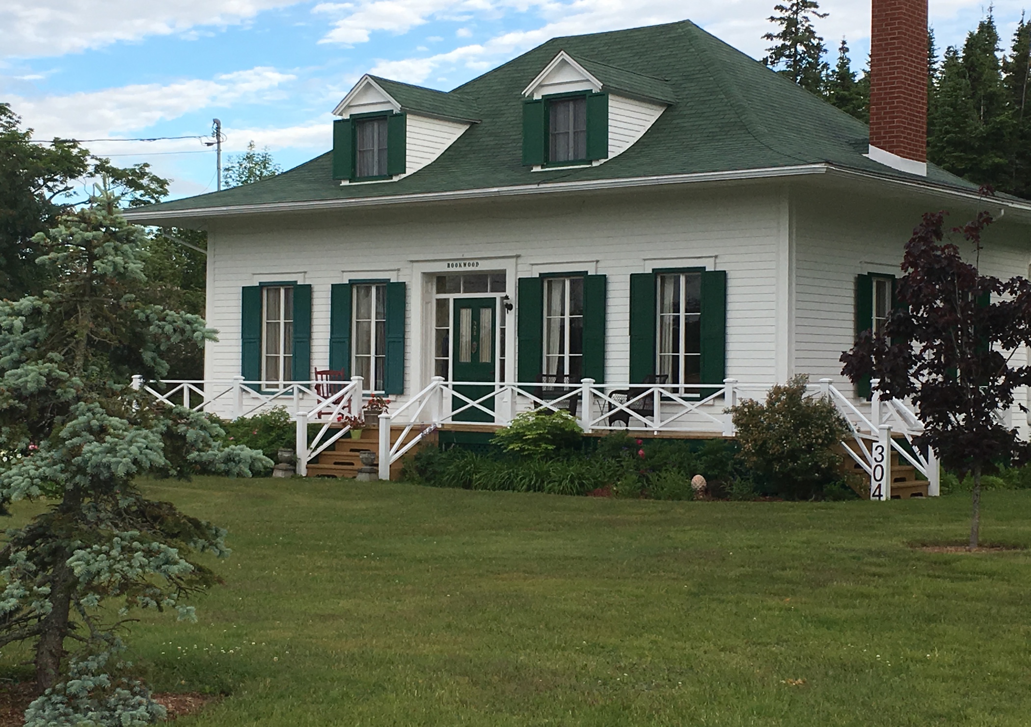 A one-and-a-half-storey summer cottage with a brick chimney and two dormer windows. It has a small but tidy front lawn and plantings of shrubs and peonies along the edge of a plain verandah. There a spruce tree in the left foreground, and in the background, a cloud-strewn but sunny sky.