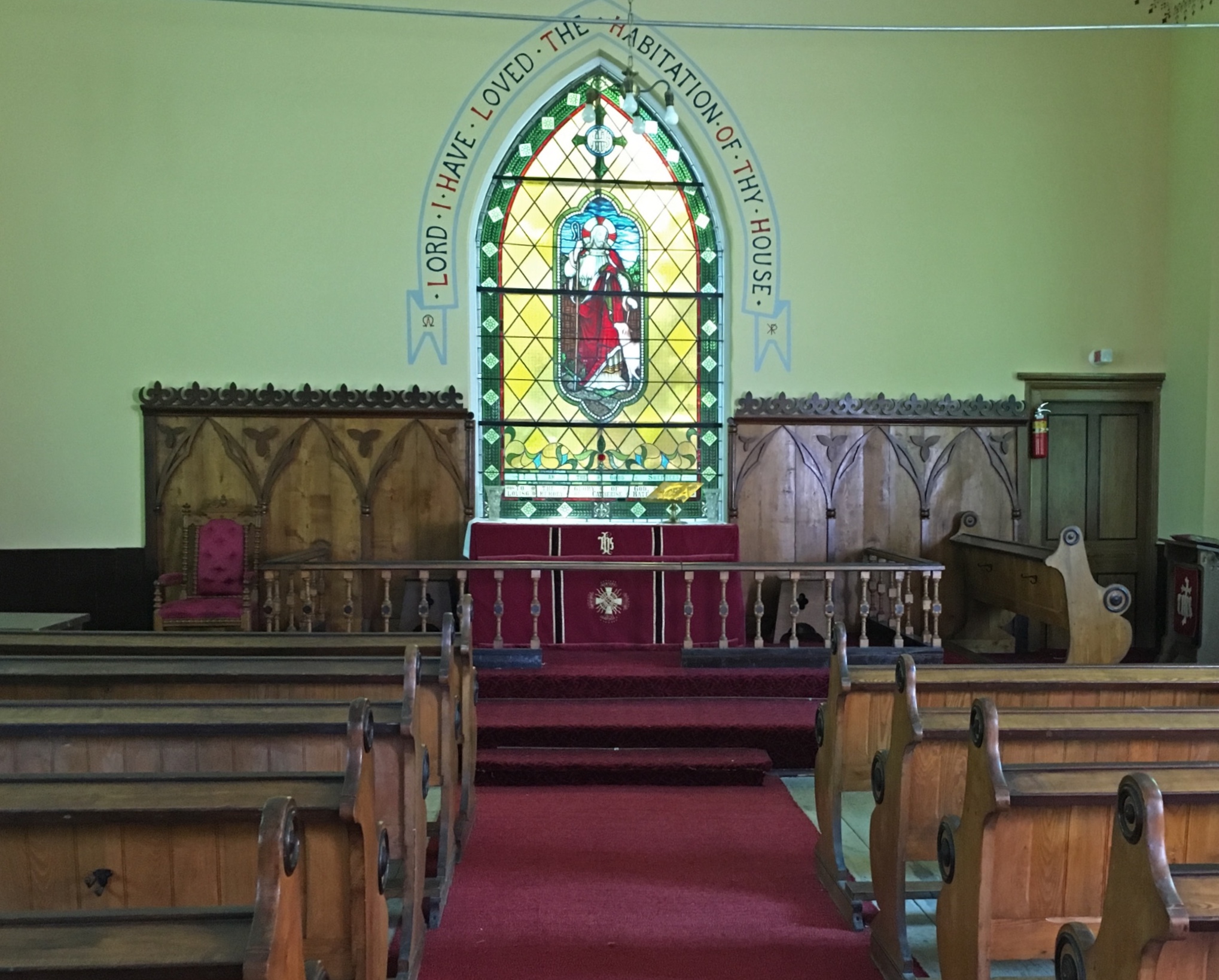 Colour photograph of St. Bartholomew's Anglican church interior, wooden pews, wooden backdrop to altar, a plain wall in the centre of which is a stained glass window.