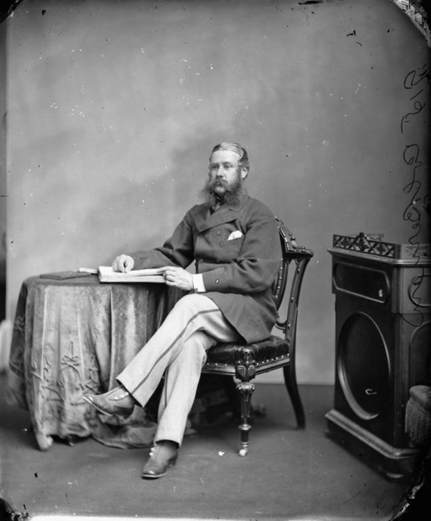 A black and white photo of Sir Hewitt Bernard in woolen short coat and striped trousers, his legs crossed, resting his right arm on a round table and a book or ledger in front of him.