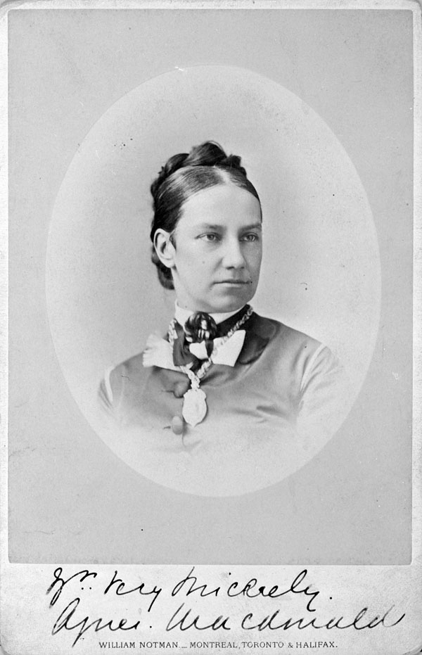 A head-and-shoulders portrait of Lady Agnes Macdonald, a young woman of about 30 to 35 years old, her dark hair pulled tightly into an arrangement of braids pinned behind her head. She wears a buttoned-up shirt, a cameo at her throat and a necklace the details of which are indistinguishable. Her expression is a mixture of thoughtful and stern.