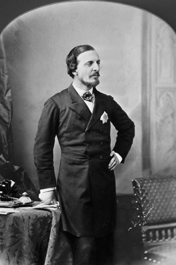 A formal black and white portrait of Lord Dufferin in a dark suite with a white handkerchief in the front pocket, standing with his left hand on his hip.
