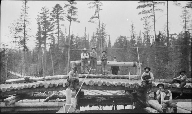 a black and white photo of nine men in overalls and shirts and hats, sitting randomly upon a wooden structure that supports a load of lumber laid in a single row; in the background stand tall trees.