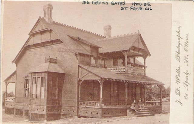 An old postcard showing a two-storey wooden house with fine details in woodwork and trim; two people sit on the front step.