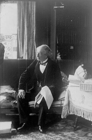 Sir John A. Macdonald in a formal suit sits upon a chesterfield holding an open sheaf of papers on his knee.