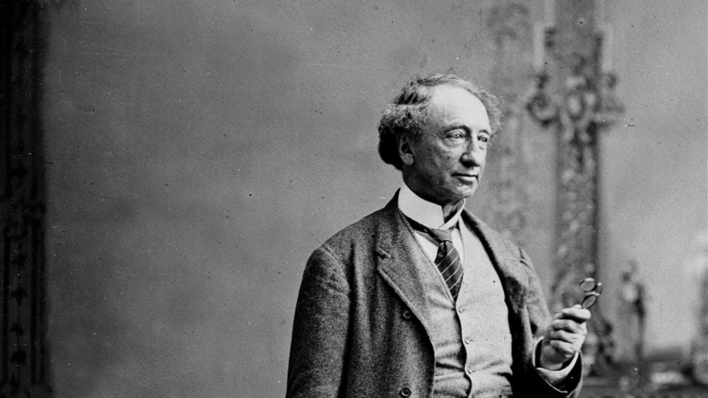 Une photo en noir et blanc de Sir John A. Macdonald, un homme âgé, vêtu d’un costume en laine, tenant dans sa main gauche une paire de lunettes.