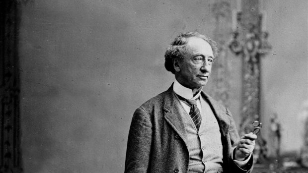 A black and white photo of Sir John A. Macdonald, an older gentleman, wearing a woolen suit holding a pair of spectacles.