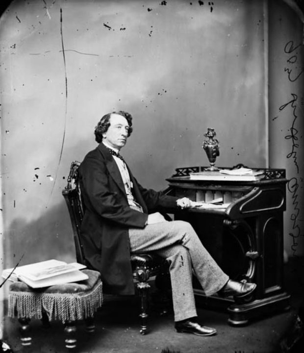 A black and white image of Sir John A. Macdonald wearing a suit, sitting at a desk, where there are books, papers and a vase.