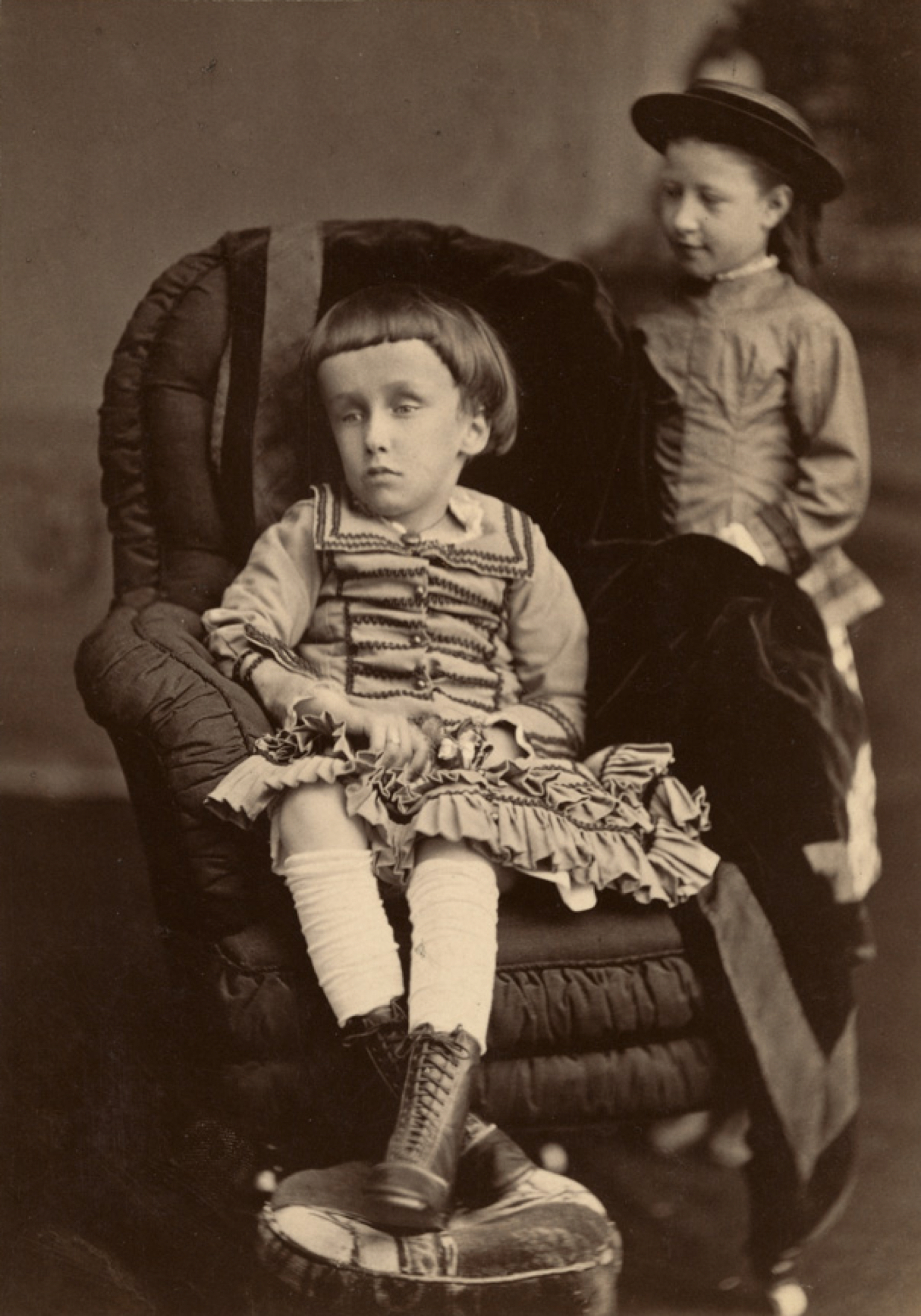 Black and white photograph of Mary Macdonald, a small child, propped up in a chair with her legs crossed in front of her, with a vacant expression on her face. Her head is abnormally large and she has dark hair combed down in bangs over her forehead.