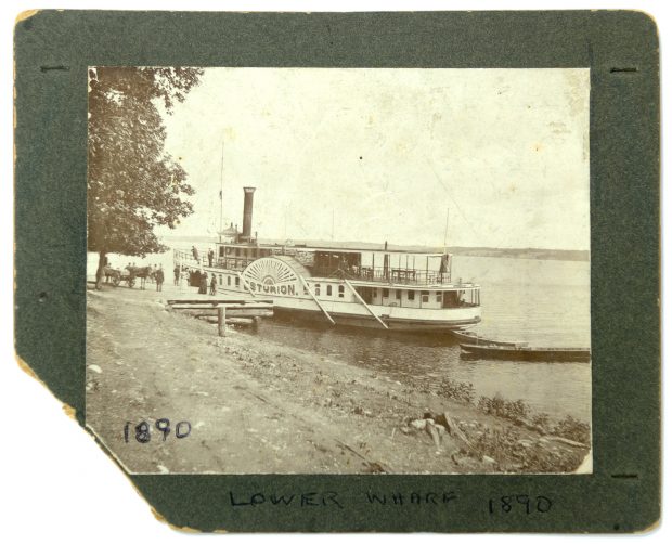 Image of large steamboat alongside dock, with people and horse-and-wagon on dock between a large tree and the boat.