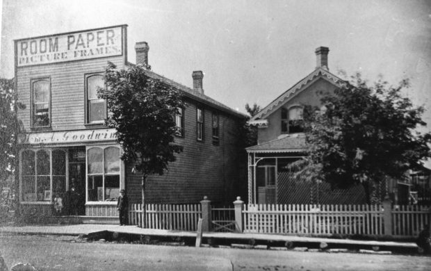 Black-and-white image of a large frame building at a street corner surrounded by trees.