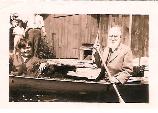Young people in a canoe being paddled by an older man. Dock and boathouse visible in the distance.