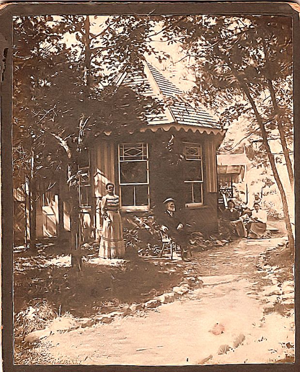 Image of a man in a straw hat sitting cross-legged in front of a cottage surrounded by tall trees, with a woman standing to his right and other figures seated in background.