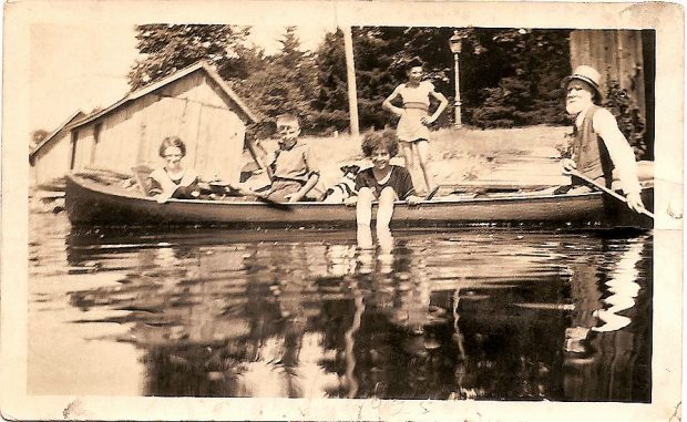 Young people in a canoe being paddled by an older man. Dock and boathouse visible in the distance.
