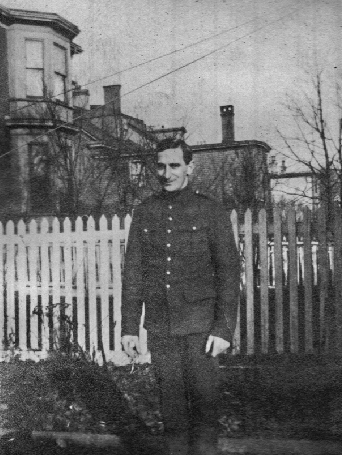 Black and white archival photograph of a man in uniform standing in front of white picket fence in front of homes, trees, and wire lines.