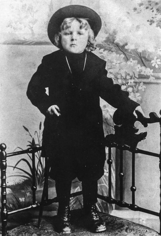 Black and white archival photograph of boy wearing dark outfit and a wide brimmed hat standing by a railing in photography studio.