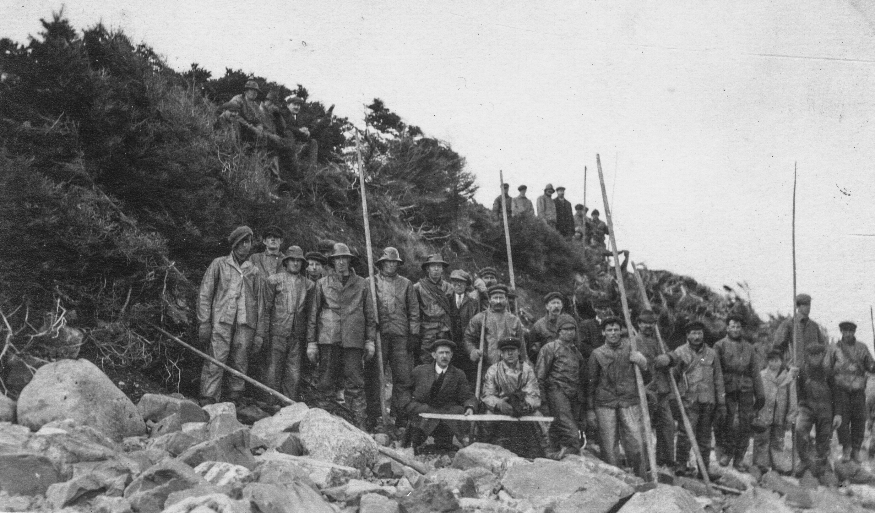 Black and white archival photograph. Group of twenty men on a beach wearing rain gear, caps, and suits holding long poles. On the hill behind the group of men are two groups of men to the left is four men sitting on the hill, and to the right is seven men standing up.