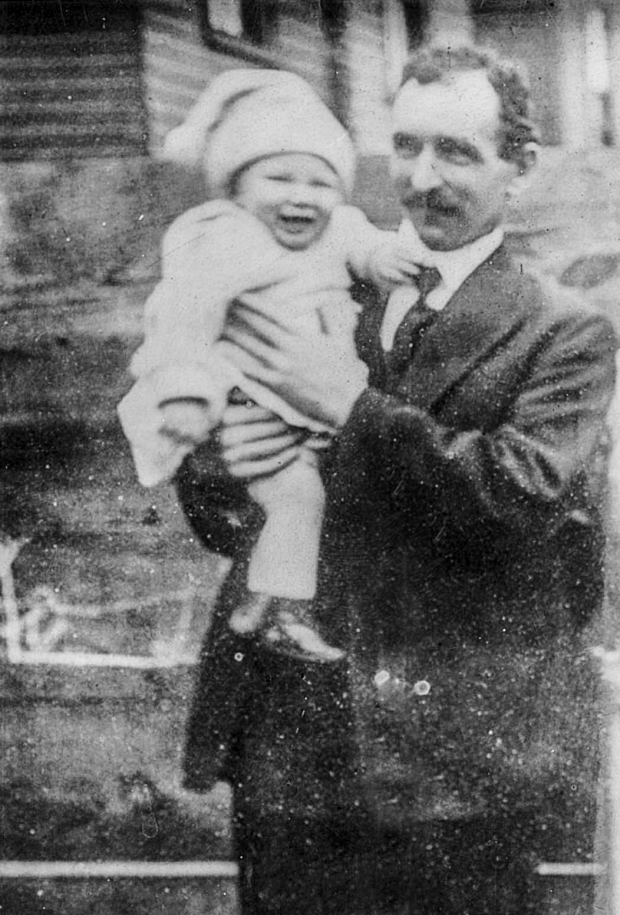 Black and white archival photograph of man standing outside of a building wearing a suit and tie and holding a baby wearing a light sweater, pants, and hat.