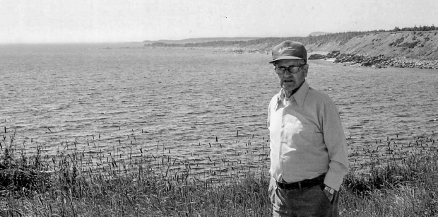 Black and white photograph of older man wearing eyeglasses, a baseball cap, a light shirt, and darker pants. Man in standing in field of grass in front of ocean landscape with hills in the background to the right.