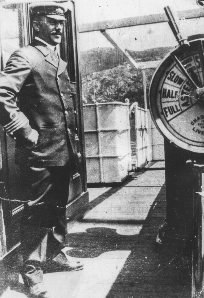 Black and white archival photograph of man in uniform wearing a peaked cap standing on the deck of a ship.