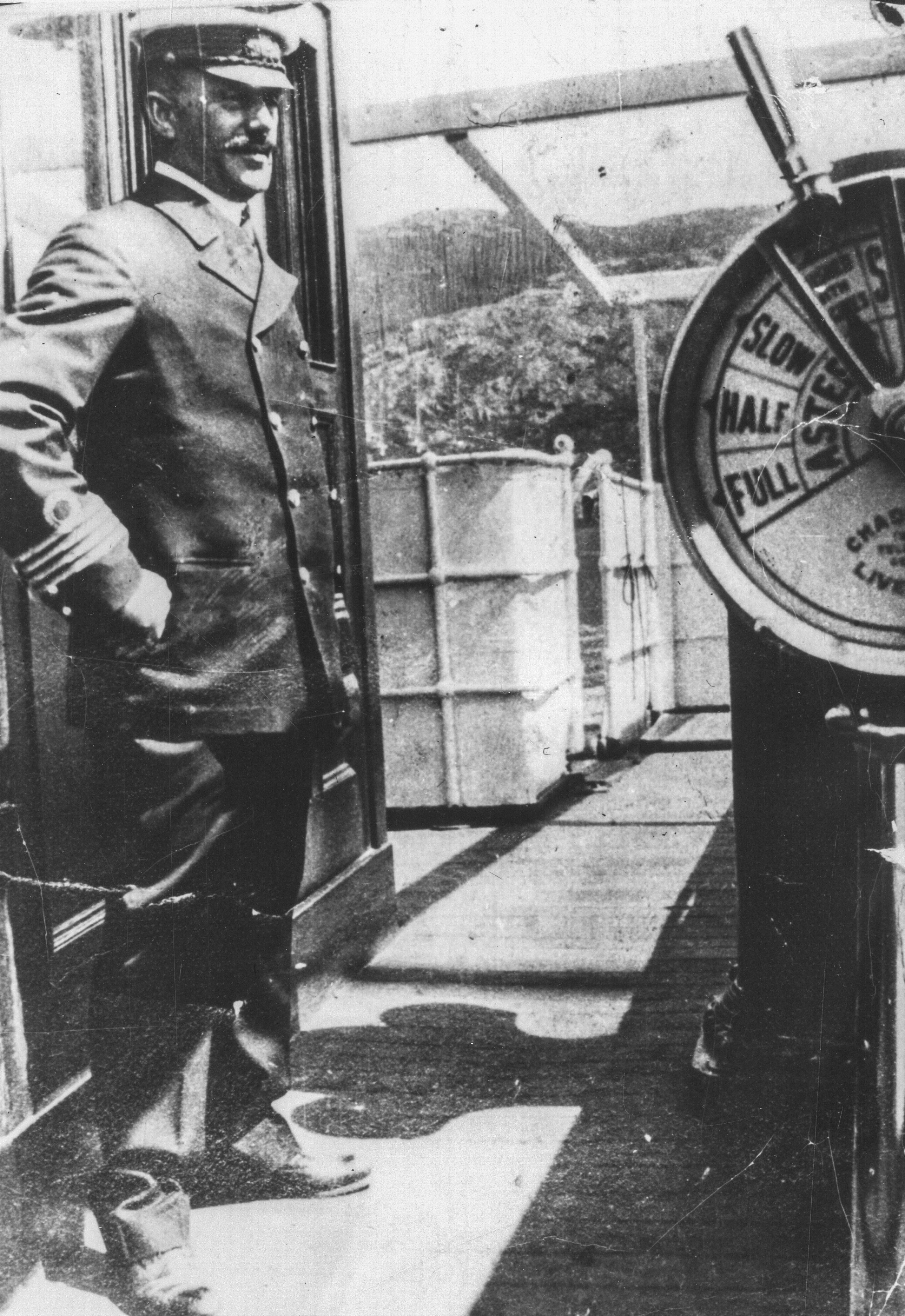 Black and white archival photograph of man in uniform wearing a peaked cap standing on the deck of a ship.