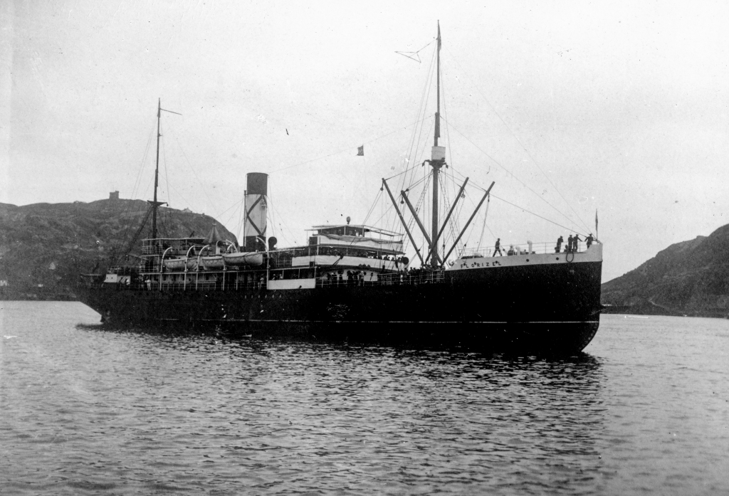 Black and white archival photograph. Large passenger liner ship, the SS Florizel, in the St. John’s harbour. Behind ship to the left is a hill with a stone tower on it, and to the right are hills. People can be seen on the ship deck.
