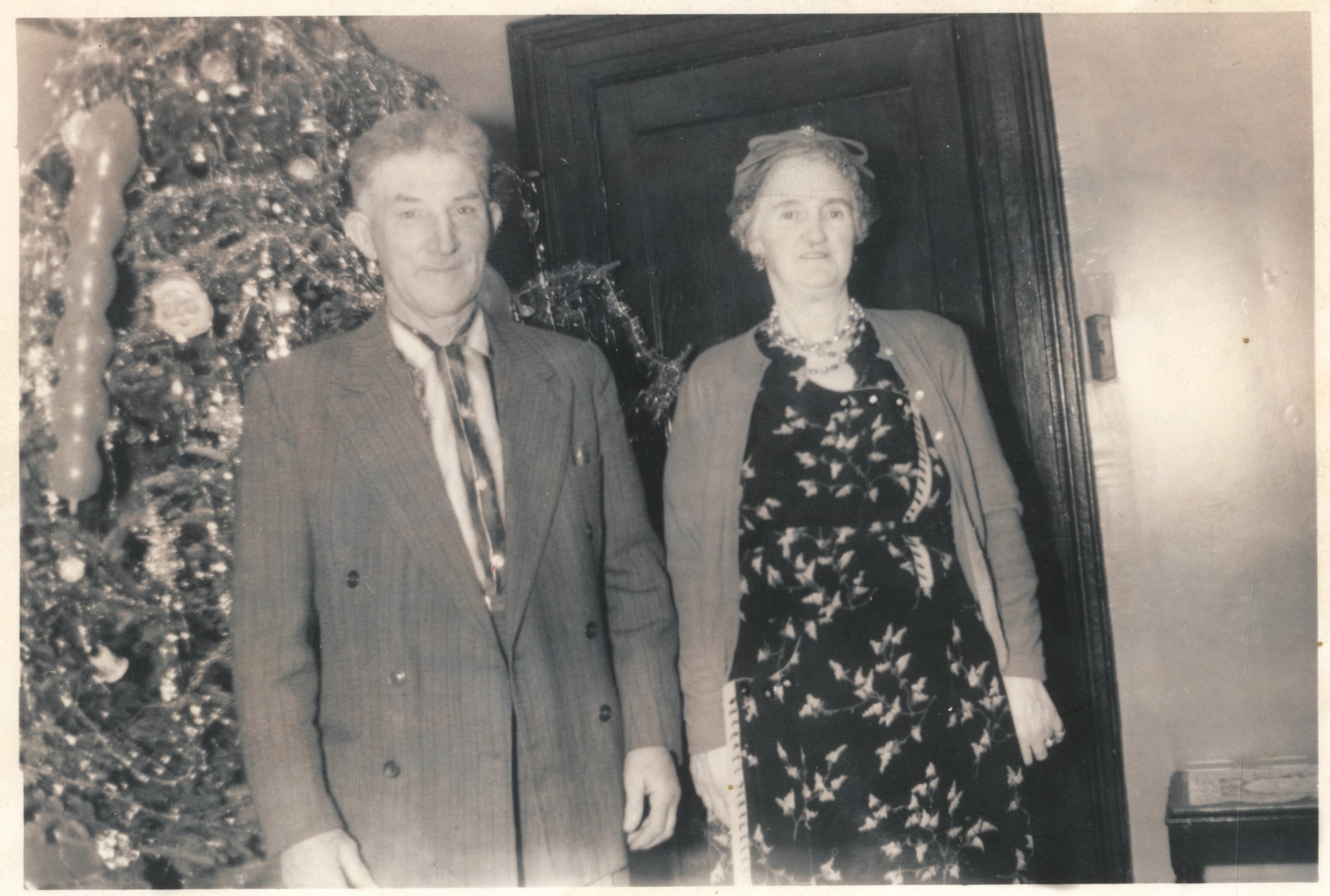 Black and white archival photograph of a man in a suit, and a woman in a dress, cardigan, hat, and necklace standing in front of Christmas tree covered in tinsel and ornaments.