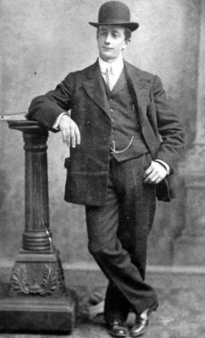 Black and white archival photograph of man leaning against post while wearing a suit, tie, and bowler hat.