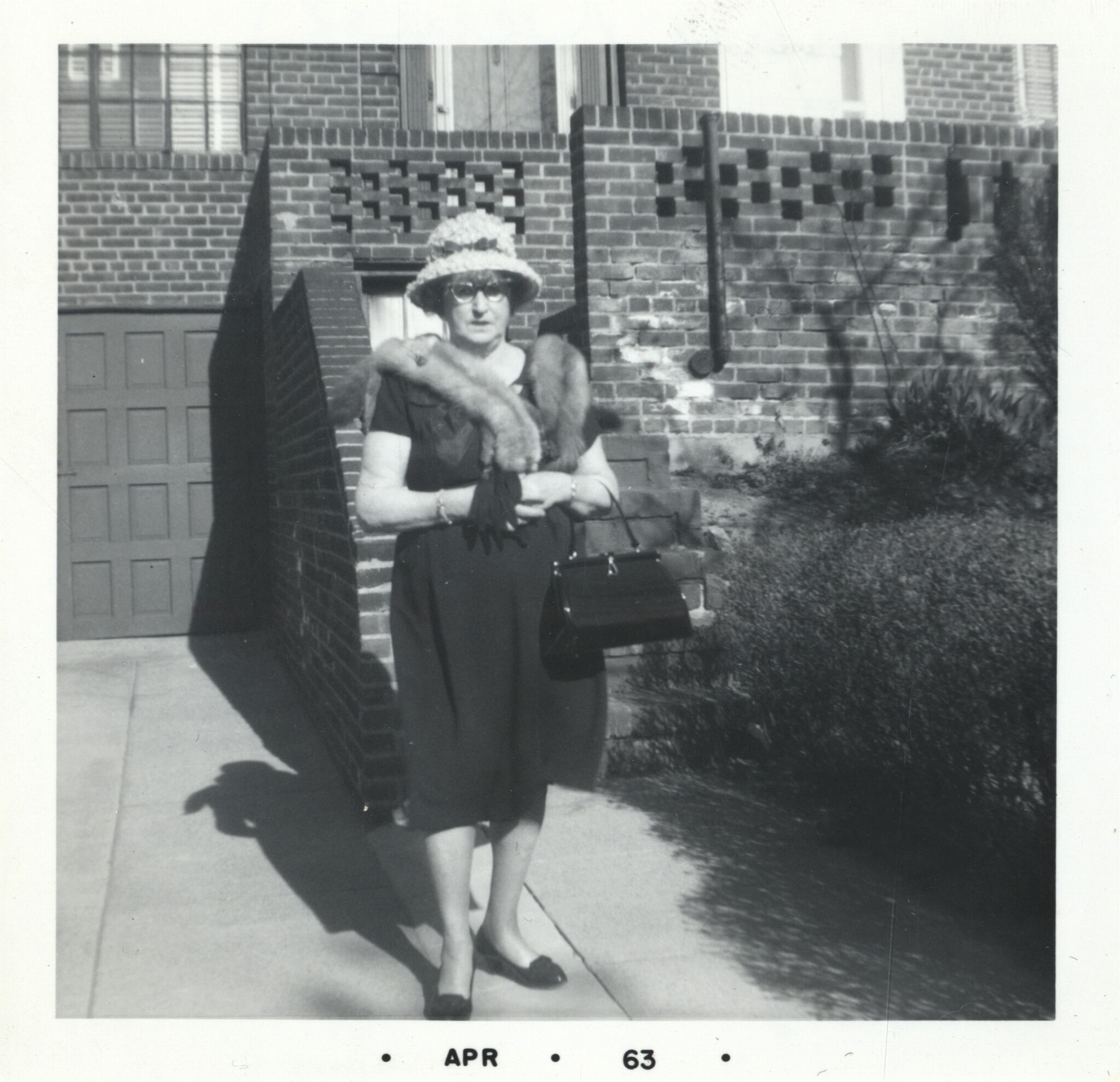 Black and white archival photograph of a woman in a dress, fur collar, hat, and glasses carrying gloves and a purse standing outside in front of a brick building.