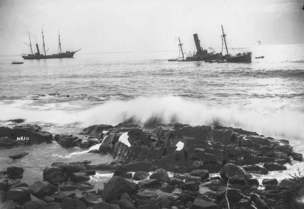 Black and white archival photograph of large passenger liner ship, the SS Florizel, run aground in the ocean just offshore from rocky beach. Seven rescue boats can be seen around the Florizel. Large rescue ship can be seen to the left of Florizel with two smaller rescue boats to the left of large ship.