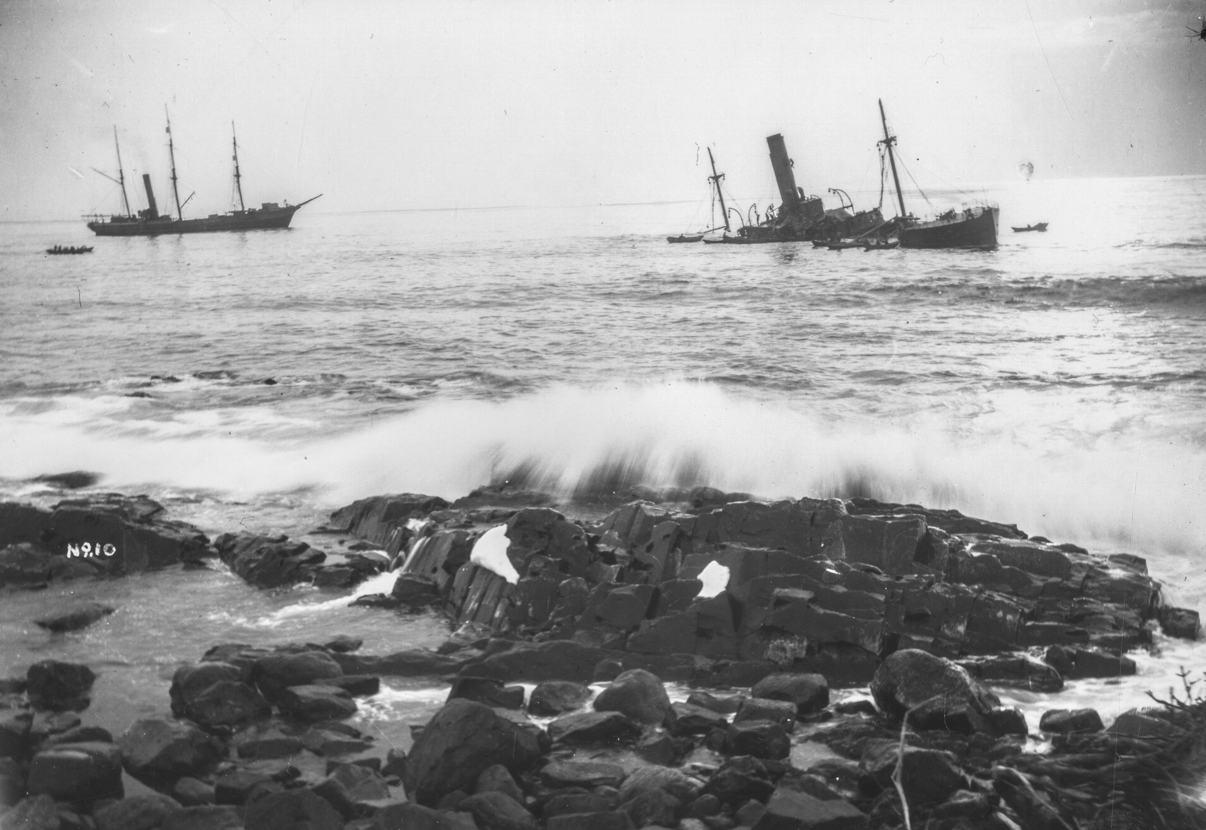 Black and white archival photograph of large passenger liner ship, the SS Florizel, run aground in the ocean just offshore from rocky beach. Seven rescue boats can be seen around the Florizel. Large rescue ship can be seen to the left of Florizel with two smaller rescue boats to the left of large ship.