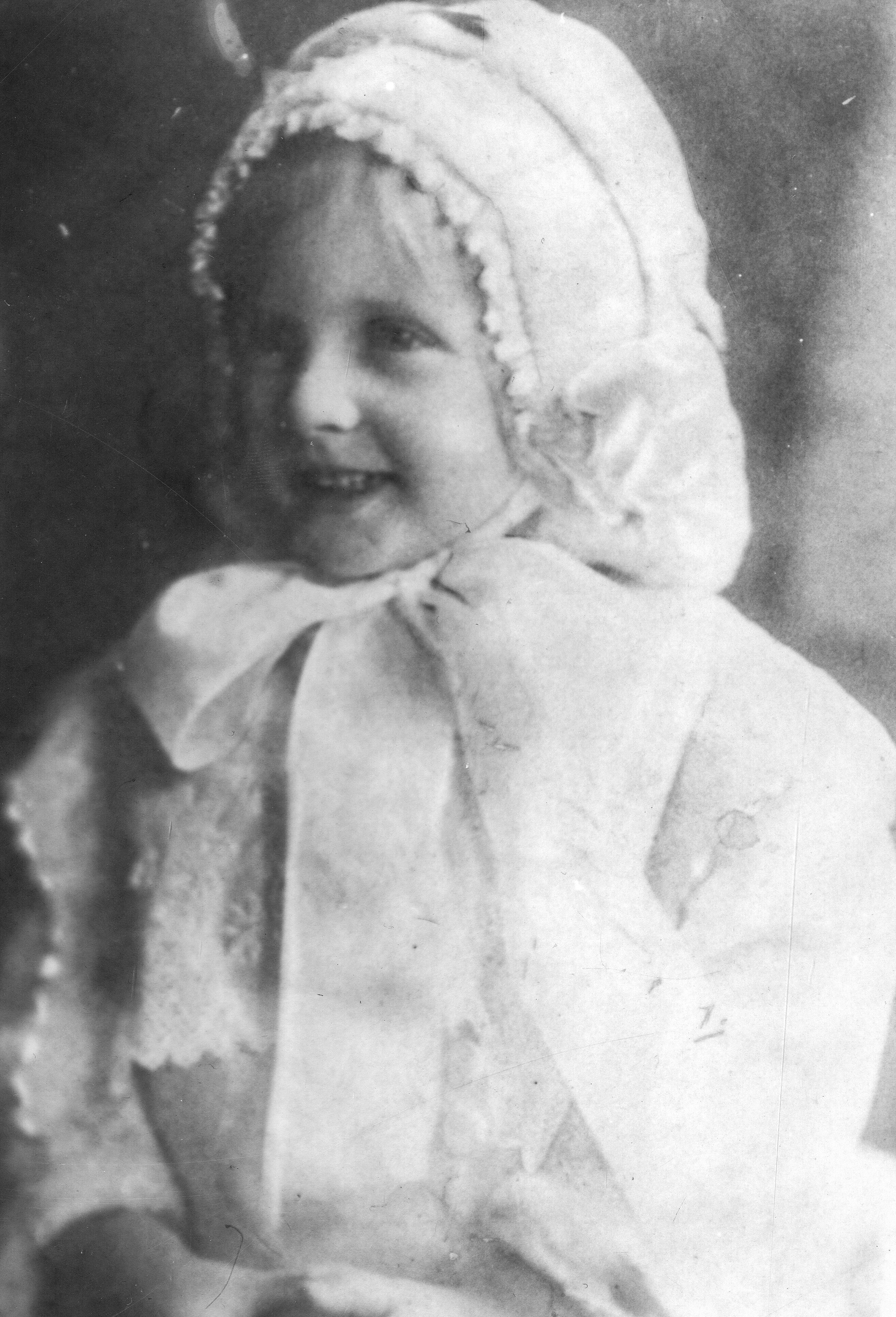 Black and white archival photograph of a girl wearing a white bonnet tied in a bow under her chin and a white dress.