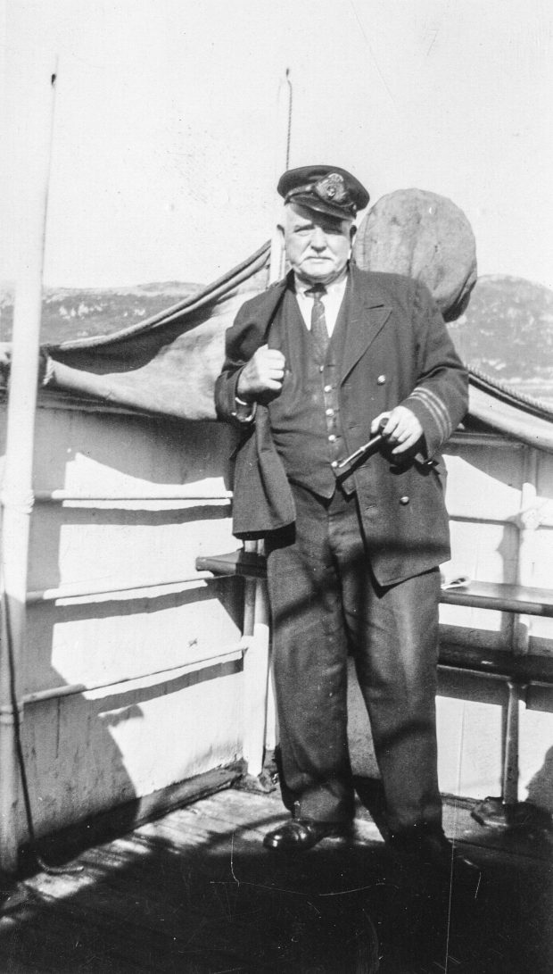 A man wearing a uniform and cap stands on the deck of a ship. He holds a pipe in his left hand. There are hills in the background.