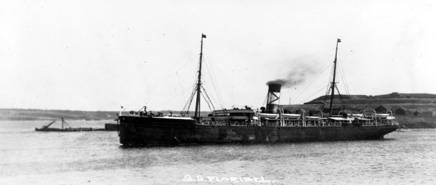 Black and white archival photograph. Large passenger liner ship, the SS Florizel, in a harbour. Behind ship to the right there are homes located on a hill, to the left there is a wharf.