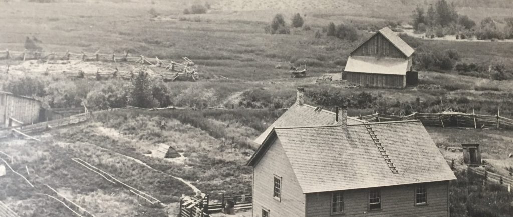 A two story wooden house is in the center of a pioneer homestead. The homestead is surrounded by marshy grass and shrubs. There are wooden fences around the house. A barn and corrals are in the background. Beyond the barn is a large hay field.