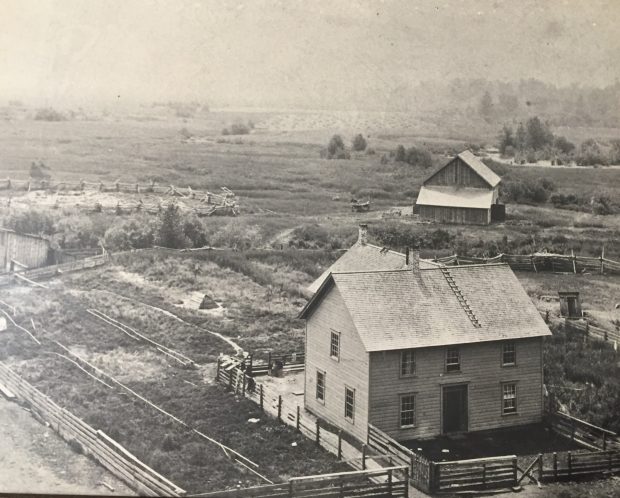 A two story wooden house is in the center of a pioneer homestead. The homestead is surrounded by marshy grass and shrubs. There are wooden fences around the house. A barn and corrals are in the background. Beyond the barn is a large hay field.