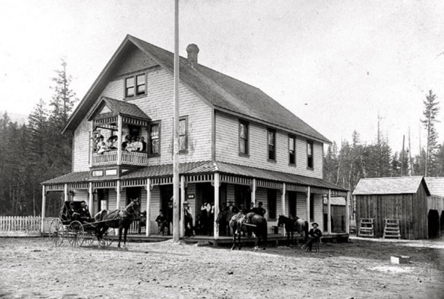 A horse and buggy and horses are standing outside a large wooden building. On the second story, several people are standing on a balcony. Several people are gathered on the covered porch. A man is sitting in a chair on the dirt street.