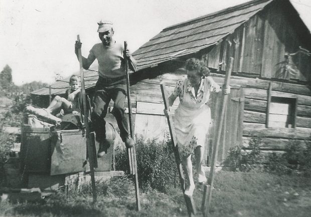A man and a woman in a dress are playing with wooden stilts. They are laughing. A boy is watching in the background. There is a wooden structure in the background that appears to be a barn.