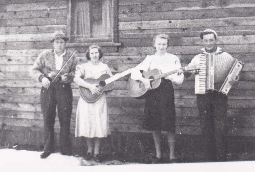 A group of four musicians stand beside a rustic wooden building. Two women have guitars. One man is holding a fiddle and the other man has an accordion.