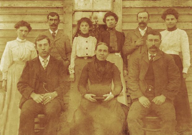 A group of pioneers are posing outside a wooden structure. Three are sitting on chairs and the rest are standing. The old woman sitting in the middle chair is holding a book.