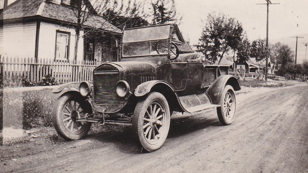 An old car with a crank start is parked on dirt city street. The car is a convertible with running boards. The street is lined with wooden houses and wooden telephone poles.