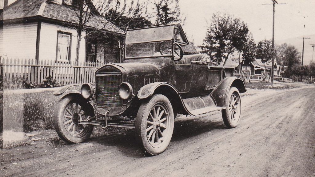 Une vieille voiture avec une manivelle est garée sur une vieille route dans une ville. La voiture est décapotable avec des marchepieds. La roue est garnie de maisons et poteau téléphoniques en bois. 