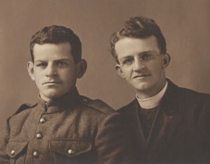 Two young men are sitting beside each other. They appear to be brothers. The man on the left has dark hair and is very serious. He is wearing at World War I army uniform. The man on the right is a priest. He is wearing round wire-rimmed glasses and has a nice smile. His hair is very wavy. He is wearing a clerical collar and dark vestment