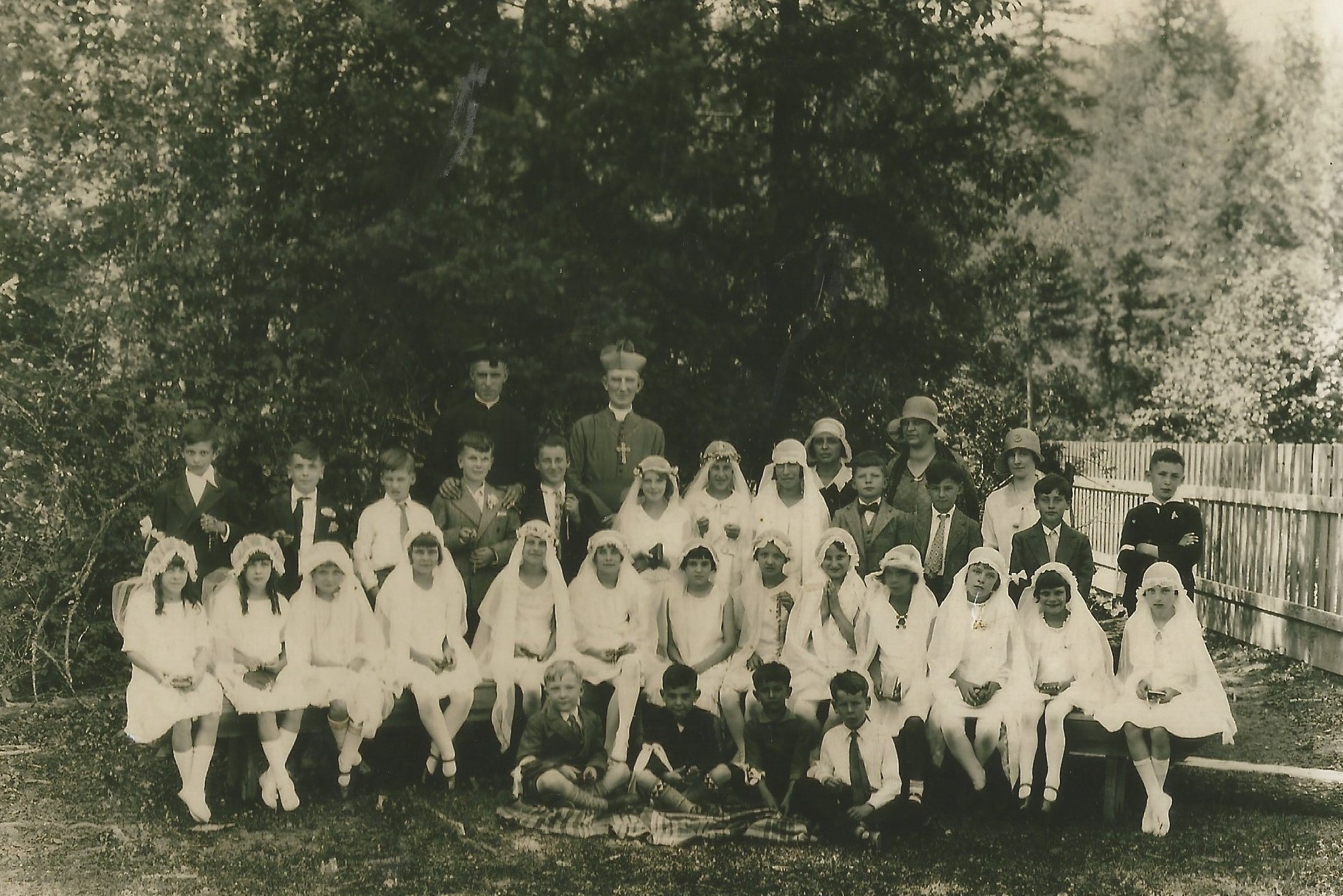 A large group of children are gathered in front of two priests. All of the young girls are wearing white dresses, white stockings, a white headdress and veil. The boys are wearing a suit and tie. Two women stand in the background.