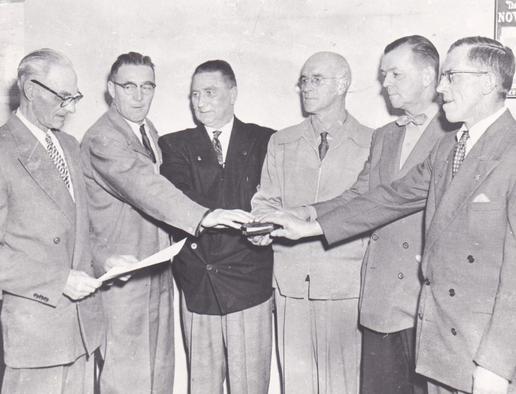 Five men all dressed in formal business suits and ties have their hands on a book. Beside them is a man reading from a paper. All of the men are very serious.
