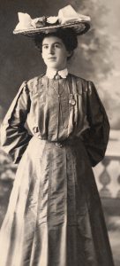 A woman is wearing a large hat with a big ribbon. She is dressed in an old fashioned long skirt and high necked blouse. The sleeves are billowy. Her hands are behind her back and she is smiling.