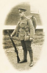 A soldier is standing on a dirt road. He has a large backpack. He is holding a rifle that is touching the ground.